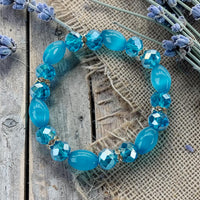 Cherry Pit Crafts blue beaded stretch bracelet with smooth and faceted beads, gold accents, and lavender sprigs.