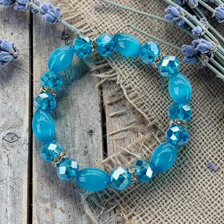 Cherry Pit Crafts blue beaded stretch bracelet with smooth and faceted beads, gold accents, and lavender sprigs.