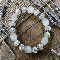 Cherry Pit Crafts white beaded stretch bracelet with faceted crystals and smooth oval beads on a wooden surface with lavender sprigs.