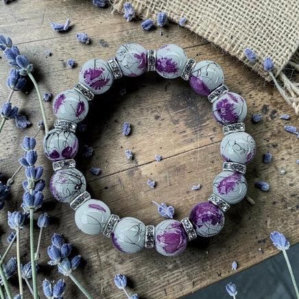 Cherry Pit Crafts stretch bracelet with white glass beads, purple ink floral details, and rhinestone accents on a wooden surface with lavender.